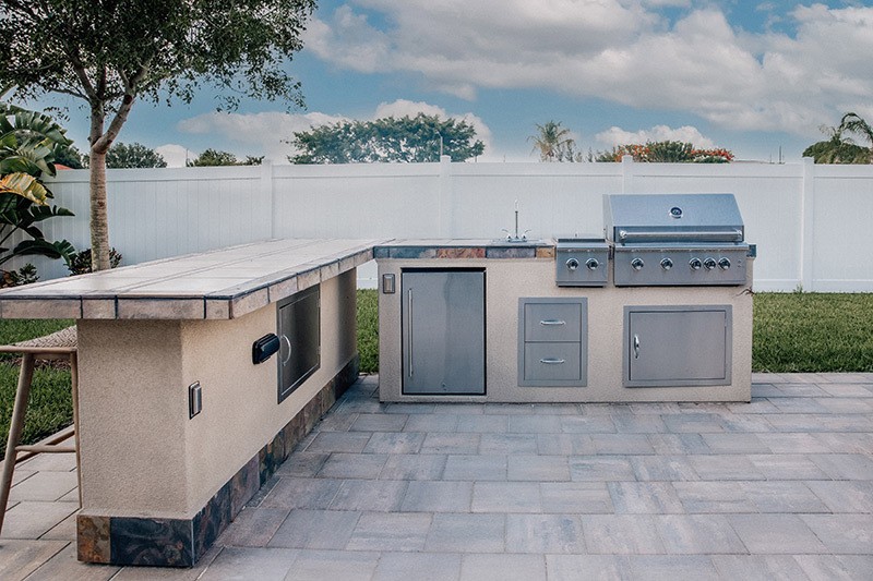 A beautiful recently constructed outdoor kitchen island featuring a stainless steel BBQ, Smoker, Sink, built in mini fridge, prep station, and long side bar for sitting.