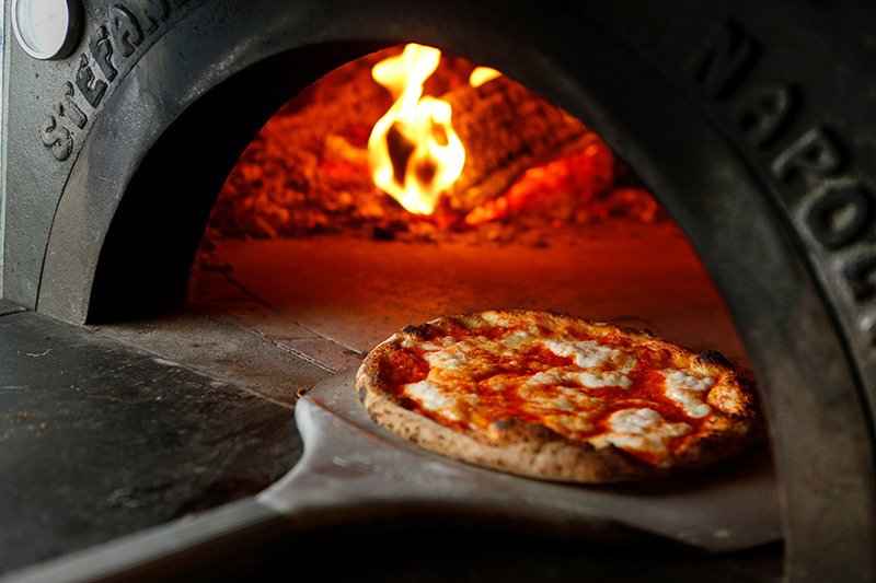 A close up images of a cheese pizza being taken out of a outdoor wood fire pizza oven.