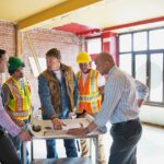 A foreman, two construction workers, and two businessmen discuss renovation plans in unfinished room