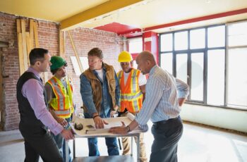 A foreman, two construction workers, and two businessmen discuss renovation plans in unfinished room