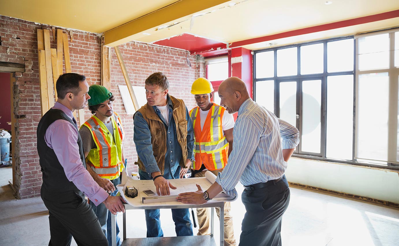 A foreman, two construction workers, and two businessmen discuss renovation plans in unfinished room