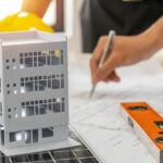 Architect model of 4-story building, hard hat, level, and hand holding pen over plans.