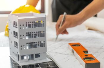 Architect model of 4-story building, hard hat, level, and hand holding pen over plans.