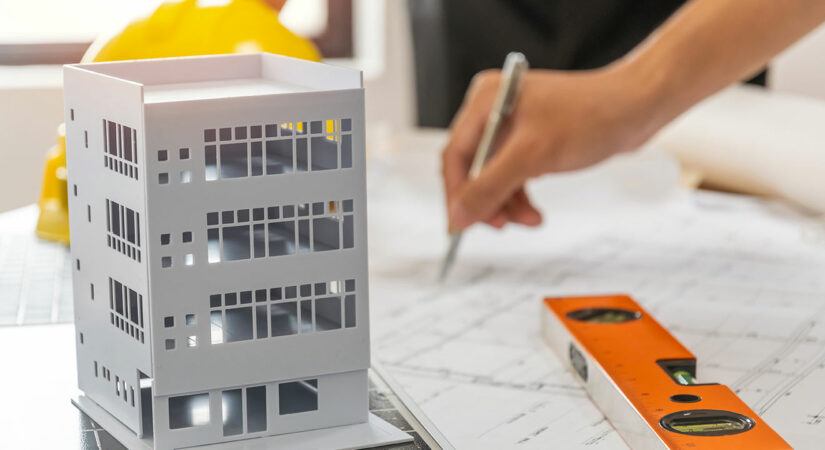 Architect model of 4-story building, hard hat, level, and hand holding pen over plans.