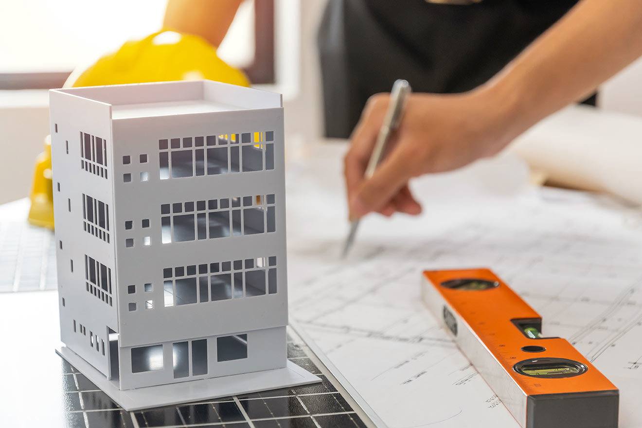 Architect model of 4-story building, hard hat, level, and hand holding pen over plans.