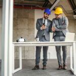 2 men in hard hats in shell of building, standing at simple desk, looking at phone.
