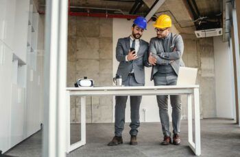 2 men in hard hats in shell of building, standing at simple desk, looking at phone.