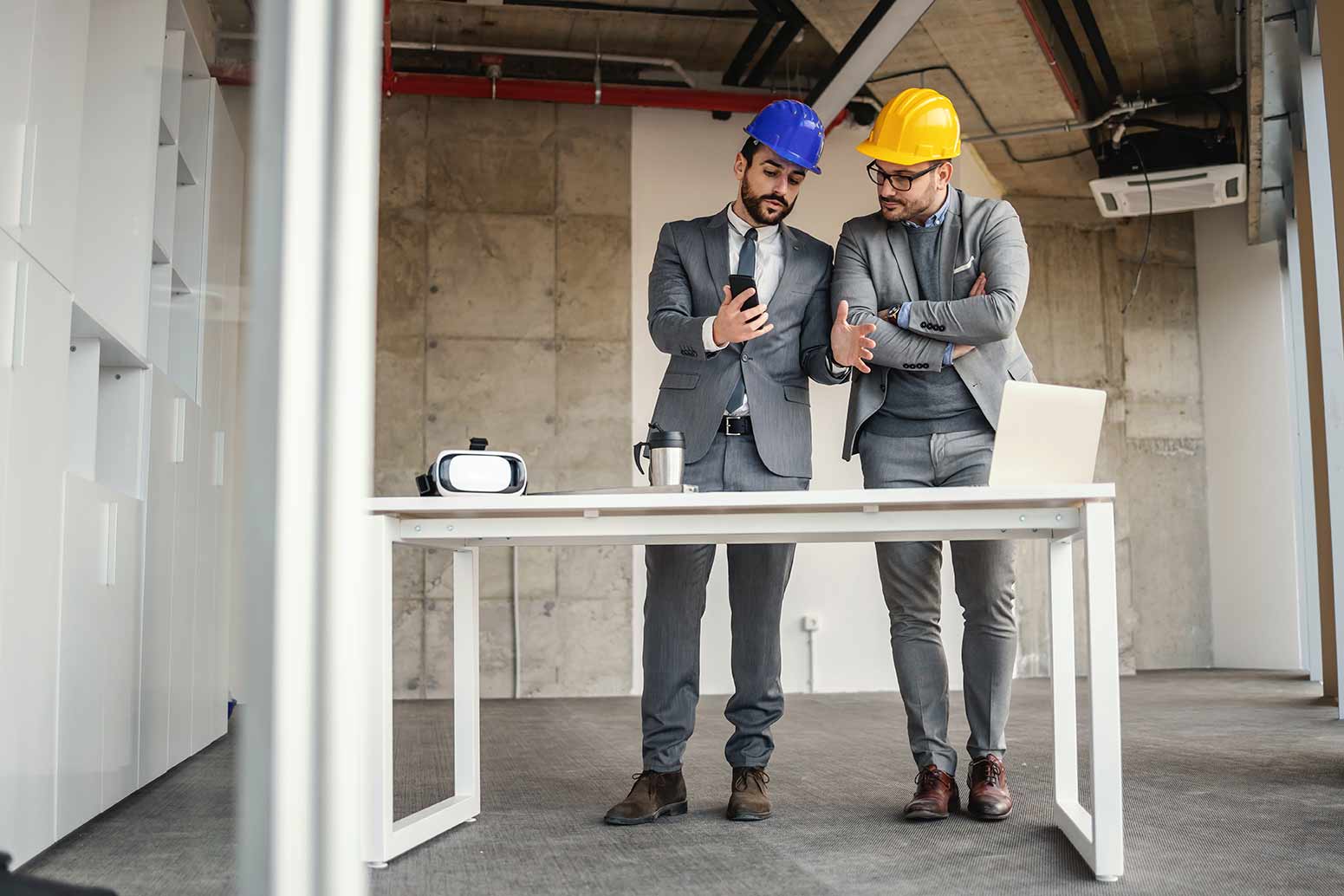 2 men in hard hats in shell of building, standing at simple desk, looking at phone.