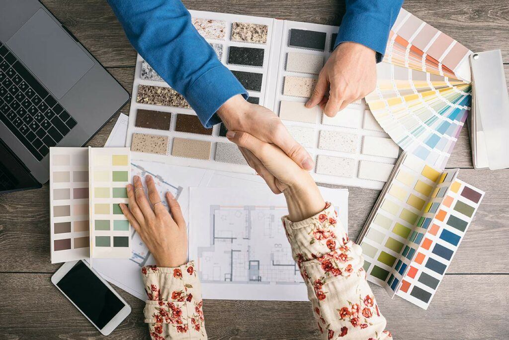 Woman shaking hands with man over table strewn with color and material swatches, plans, laptop, and cell phone