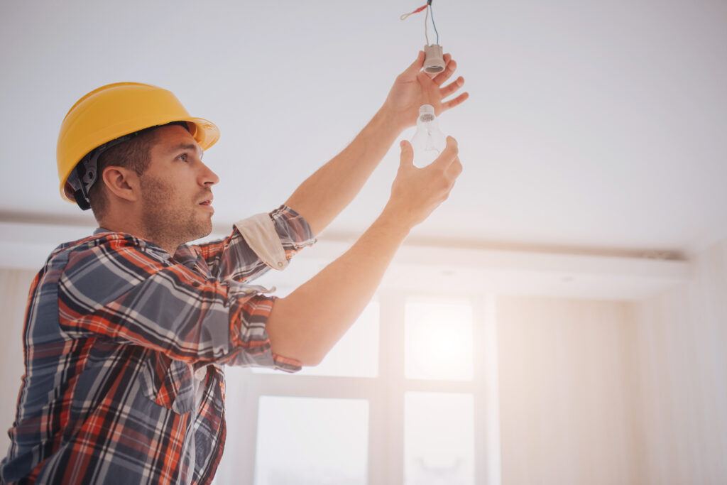A young builder in a construction hat changes a LED lightbulb for tenant improvements in an office space.