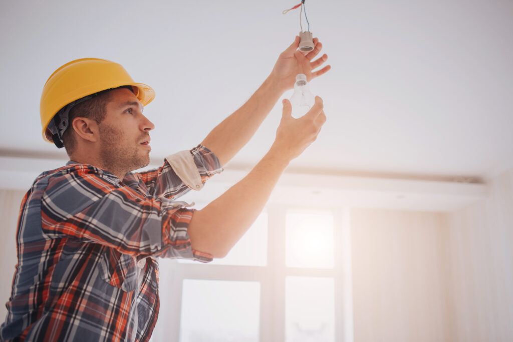 A young builder in a construction helmet changes a LED light bulb during a tenant improvement project in an office building.