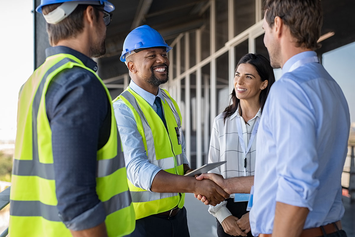 A smiling home builder in a hard hat shakes hands with a client.