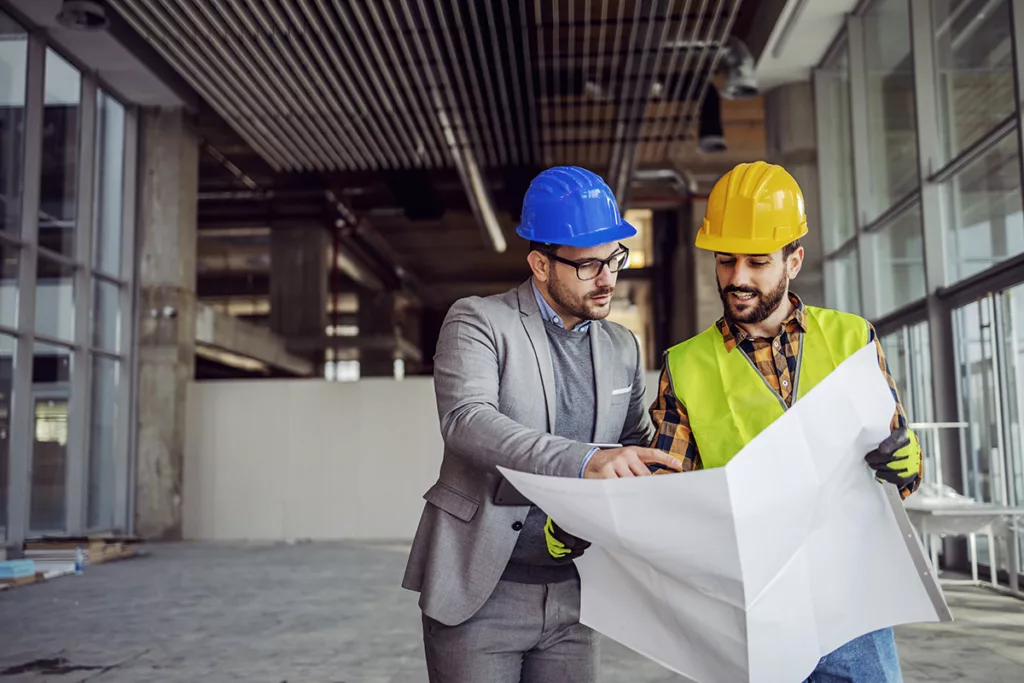 Two contractors stand in the middle of a construction site, looking over plans.