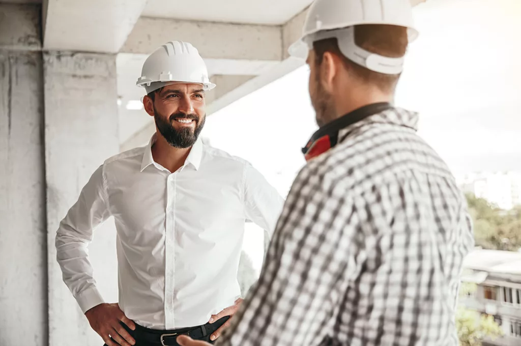 Two men wearing white hard hats facing each other and communicating with smiles