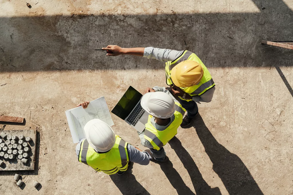 A commercial construction team stands at a construction site, looking over a computer and blueprints, wearing hard hats.