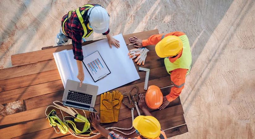 A team of commercial construction workers stand around an outdoor table, pointing at blueprints, wearing orange hard hats.