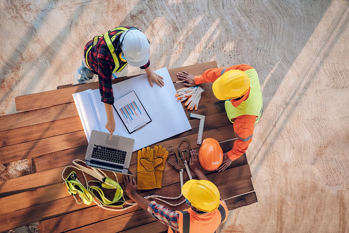 A team of commercial construction workers stand around an outdoor table, pointing at blueprints, wearing orange hard hats.