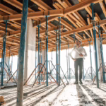 A general contractor wearing a white shirt and holding blueprints stands in the middle of a commercial building that is being constructed